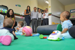 Bebês brincam no chão com tapete enquanto professora, governador e equipe observam em pé